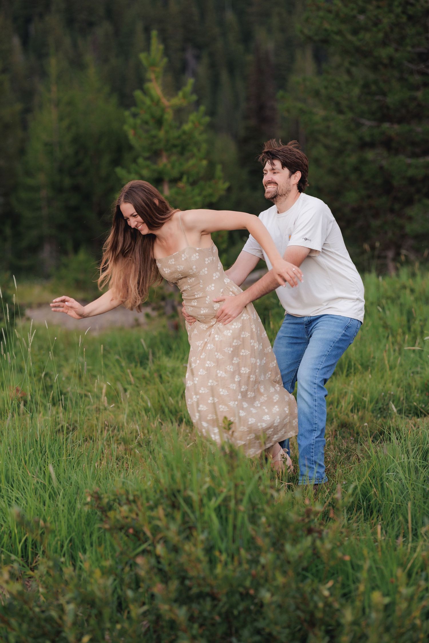 A couple dances playfully together in a grassy meadow with evergreen trees in the background.