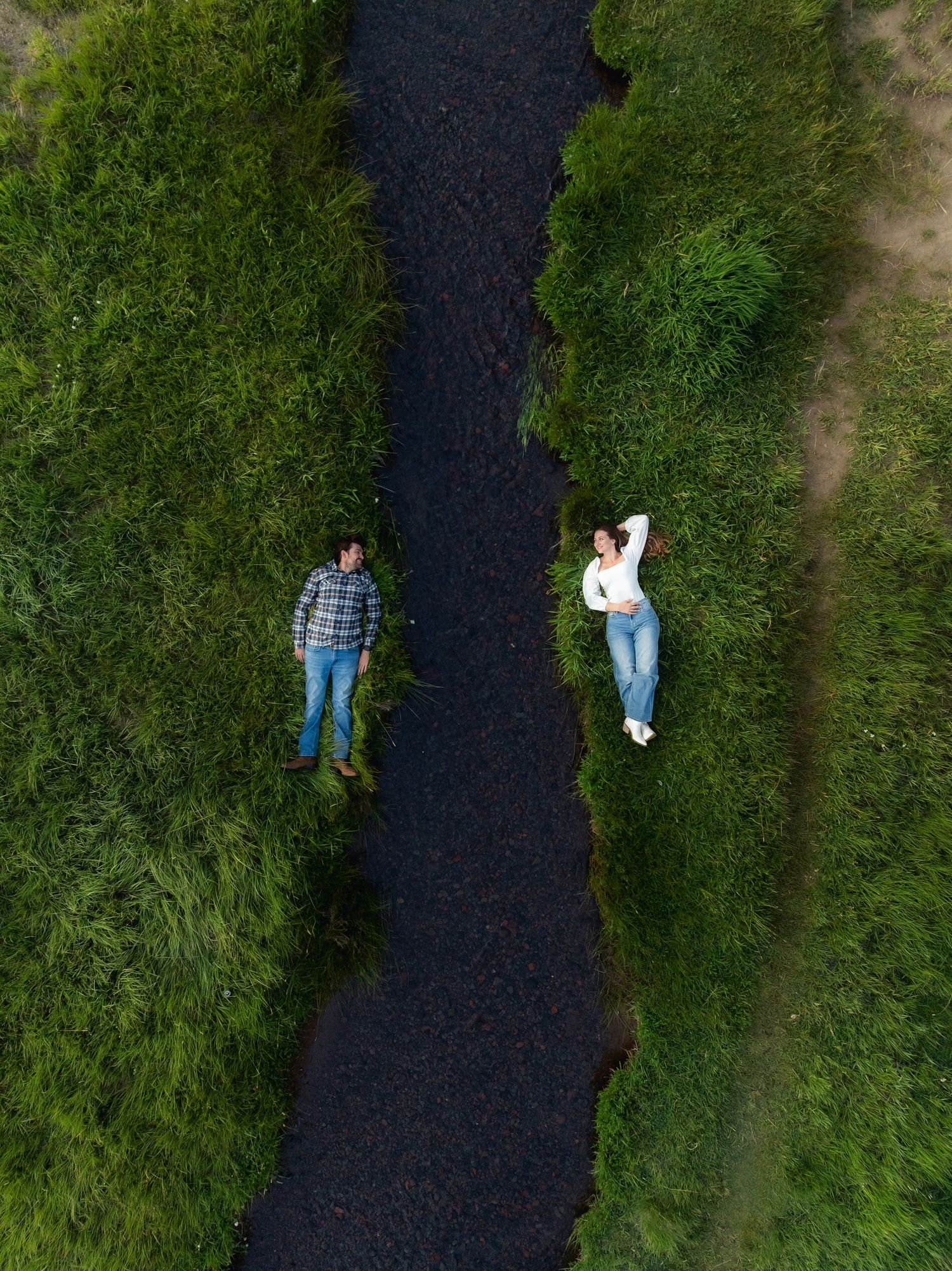 Aerial view of two people lying on opposite sides of a dark river surrounded by lush green grass.