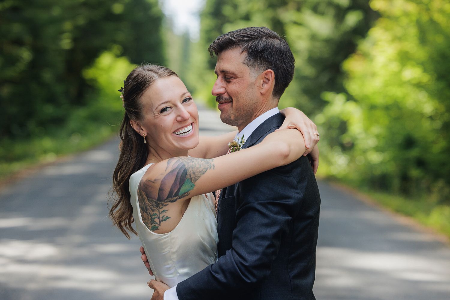A happy newlywed couple embraces and laughs together on a tree-lined path outdoors during their wedding day.