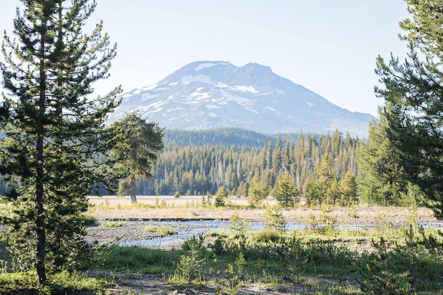 Snow-capped mountain peak rises above forested landscape with calm lake waters in the foreground.