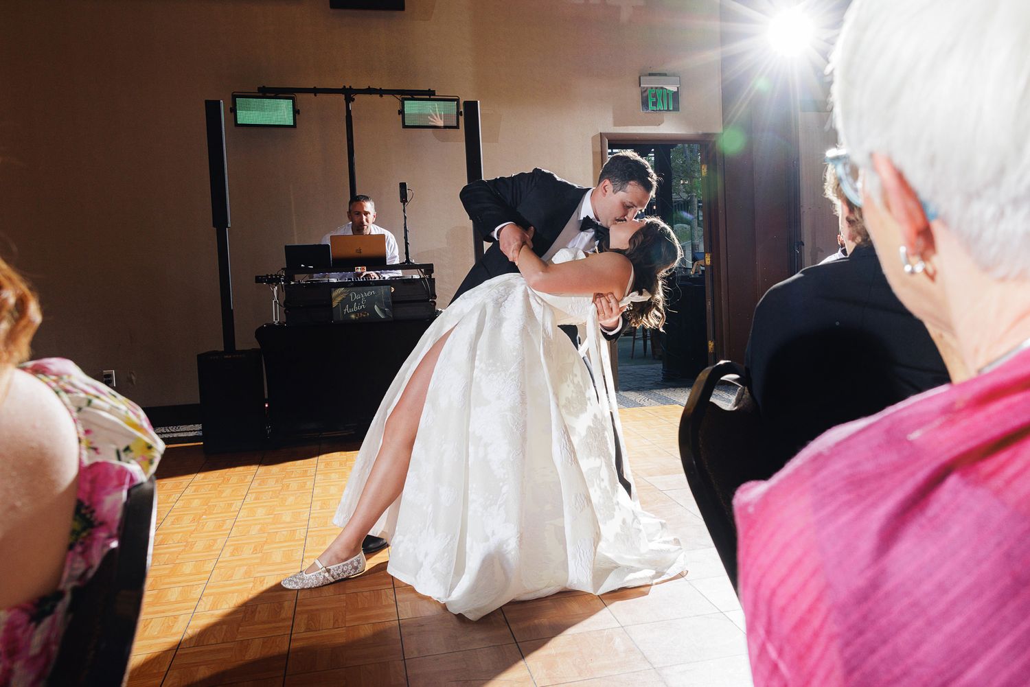 Bride and groom share romantic first dance dip at wedding reception with DJ and guests watching.