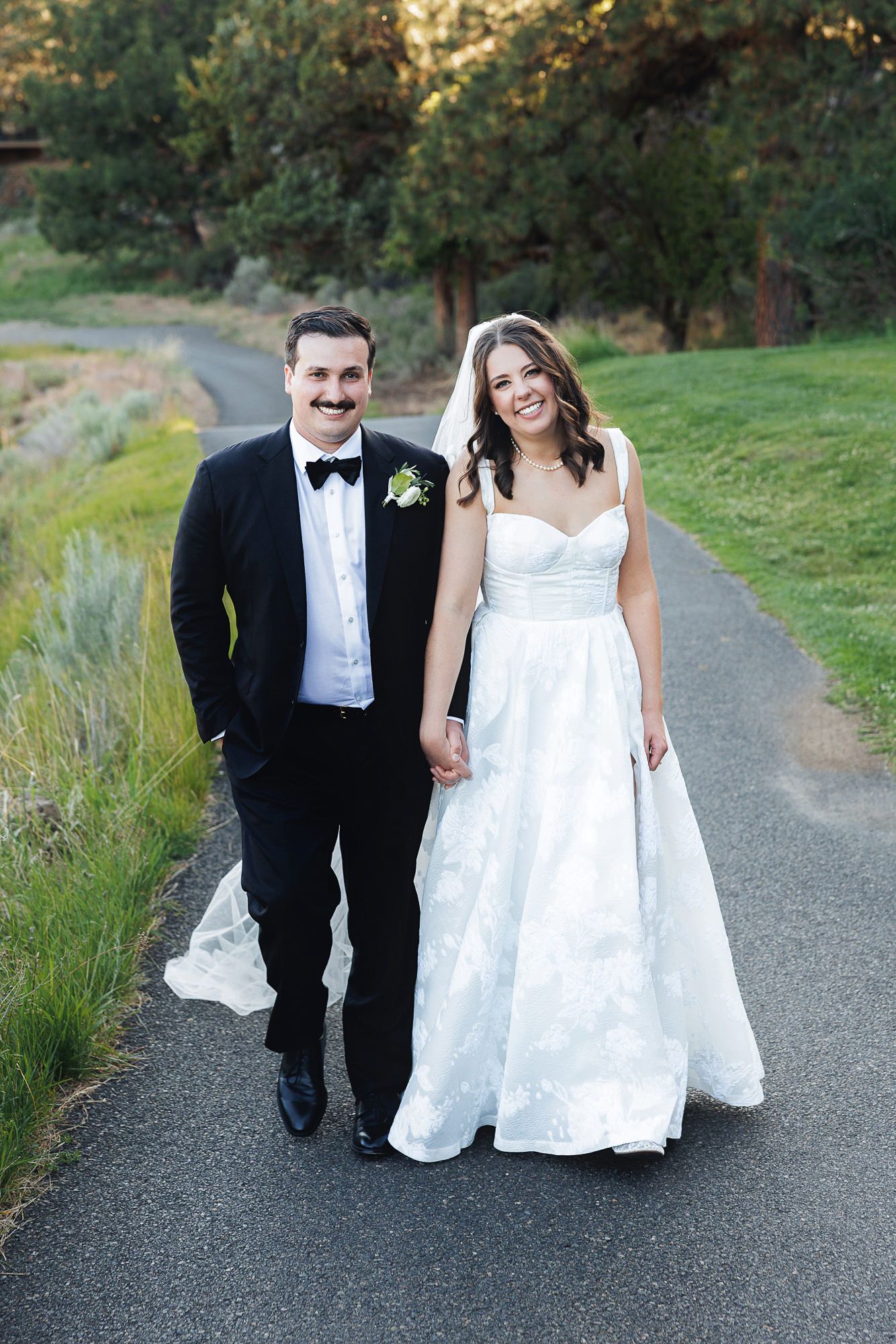 Smiling bride in a strapless floral gown and groom in a black tuxedo walk hand in hand along a path at golden hour.