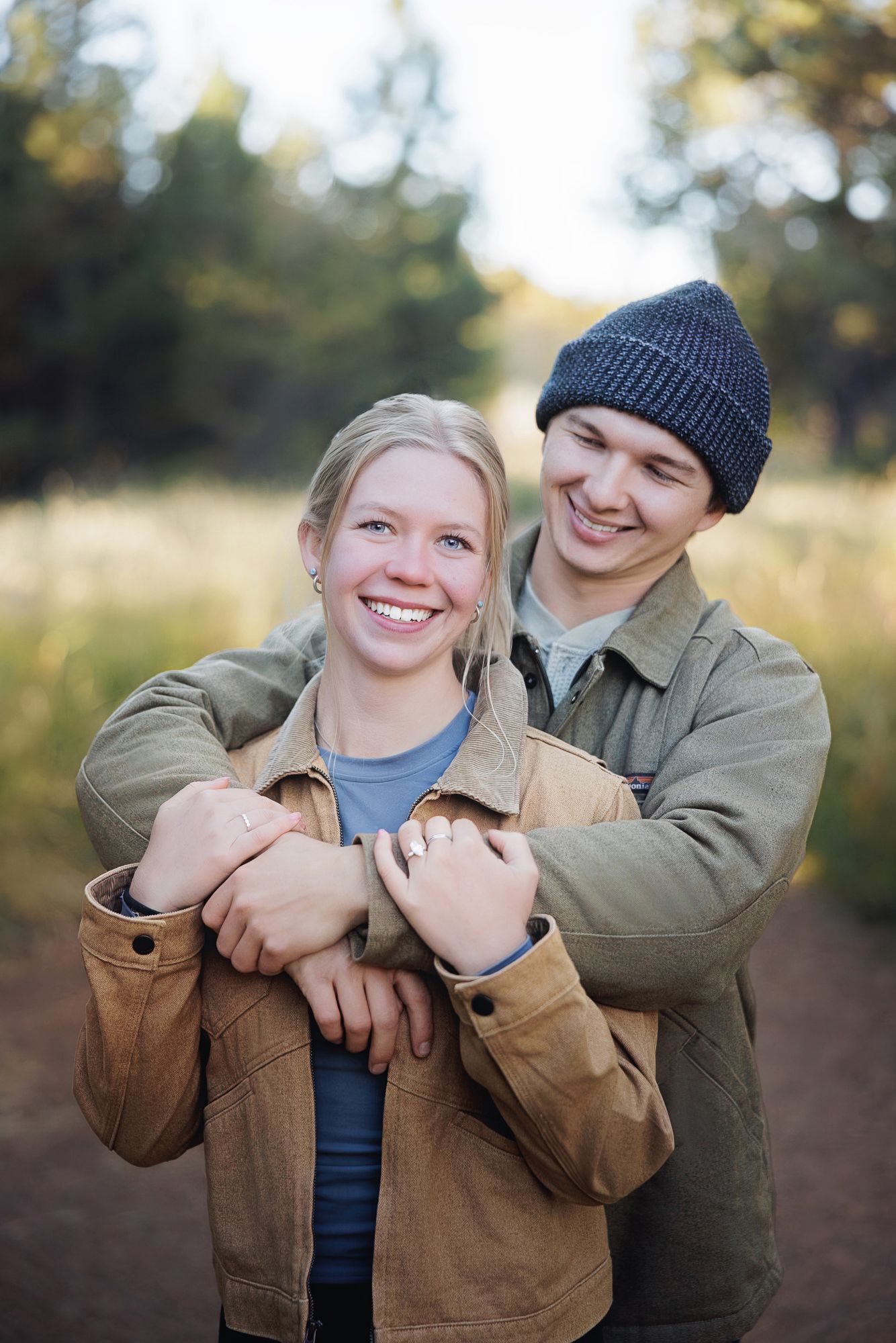 Smiling couple embracing outdoors on a forest trail, the man in a beanie hugging the woman from behind as she shows her ring.