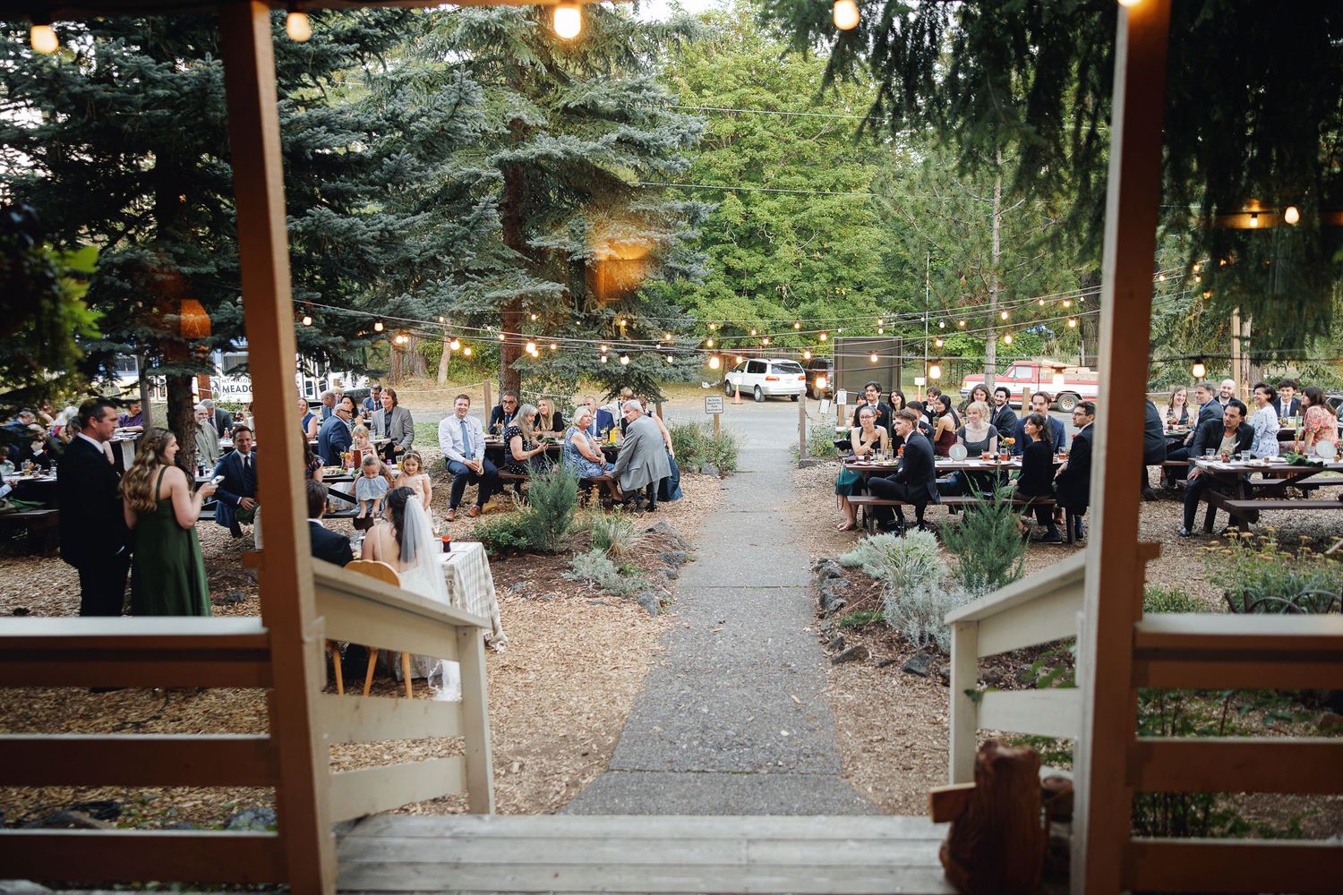 Outdoor wedding reception with string lights, guests dining at tables along a tree-lined pathway at dusk.
