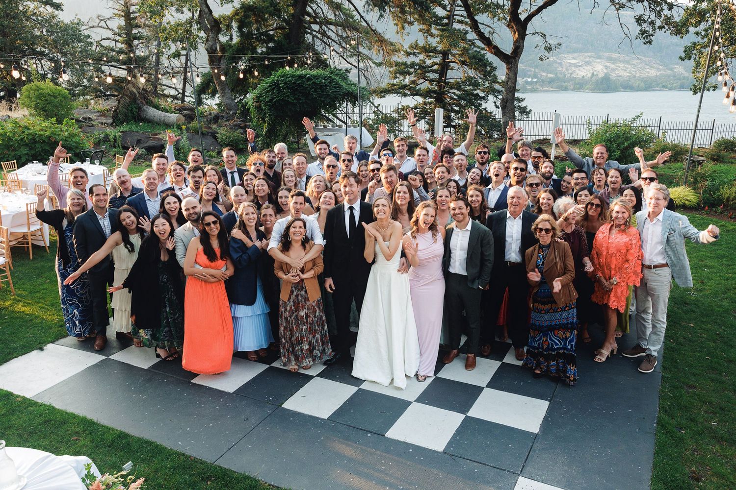 Large wedding group photo outdoors on checkered dance floor with lake view, guests in formal attire celebrating.