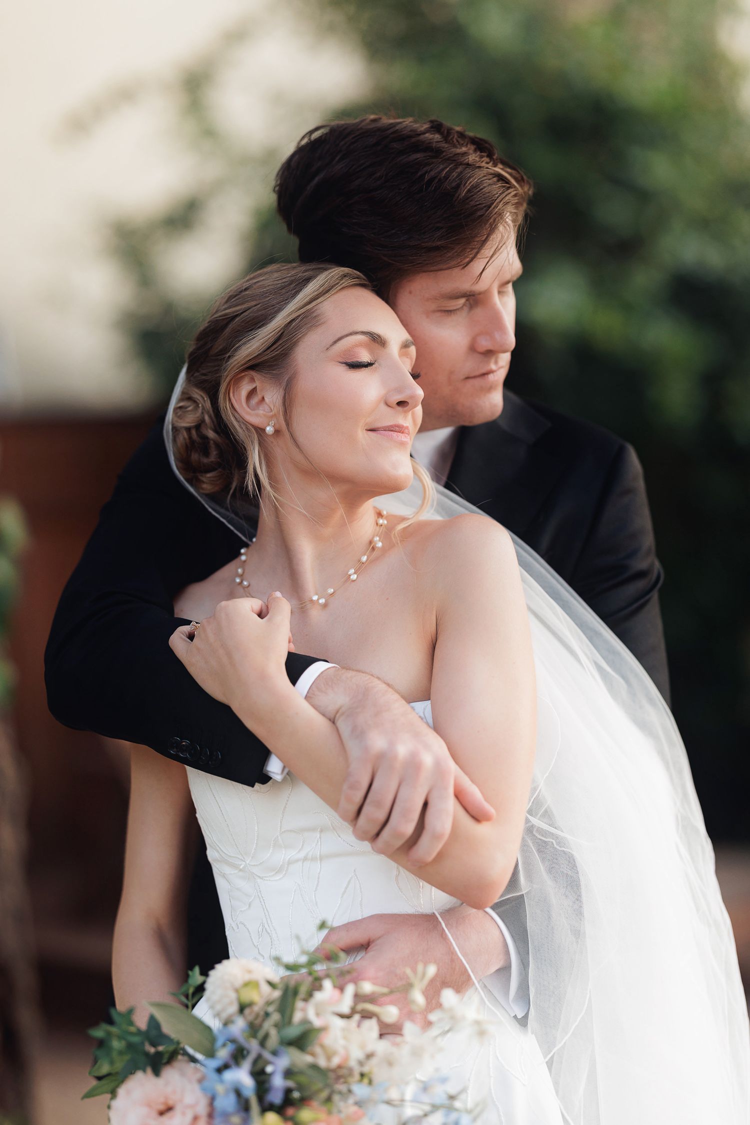 Bride and groom embracing outdoors, bride holding floral bouquet in white gown with veil, groom in black suit.