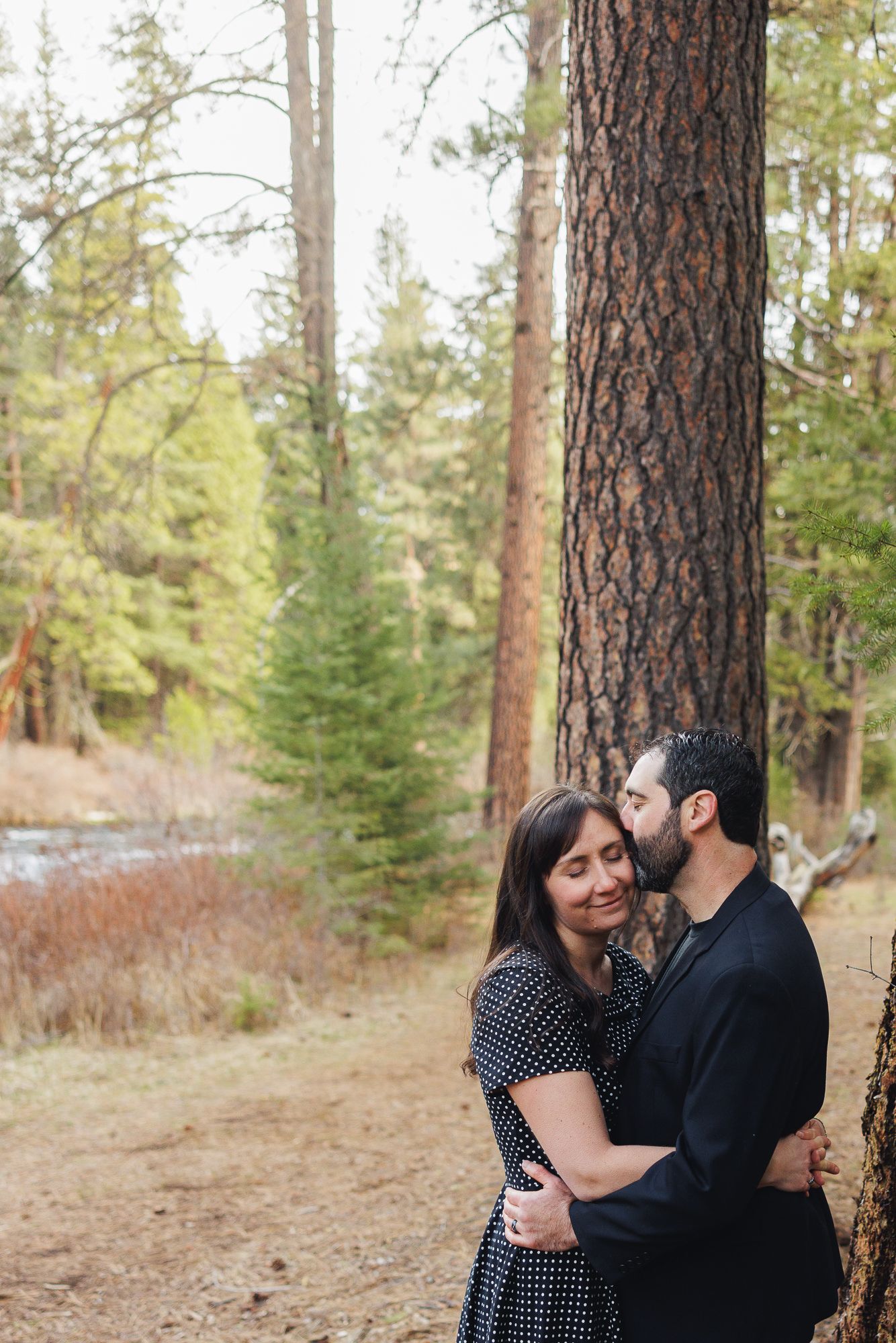 A couple embraces and shares a tender kiss in a serene forest setting with tall pine trees and a stream in the background.
