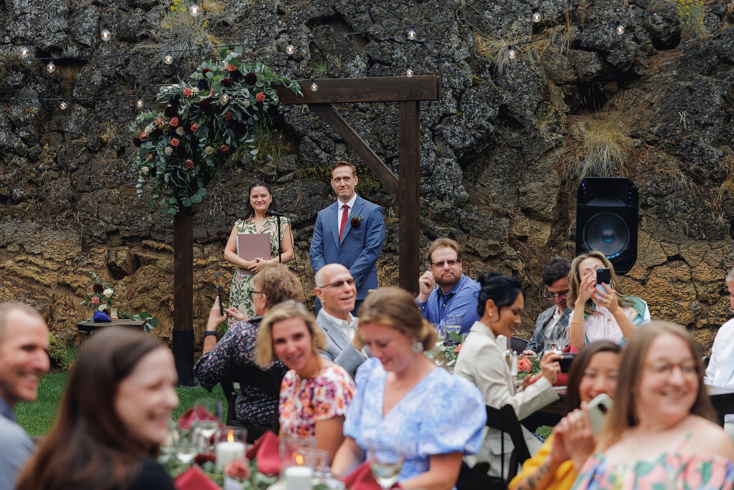 Wedding guests seated outdoors at a reception enjoy conversation and laughter in a rustic garden setting.