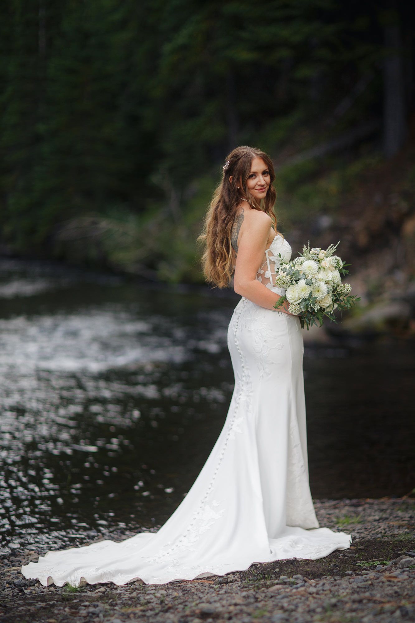 Bride in white lace wedding dress sits gracefully by a serene river holding a white floral bouquet.