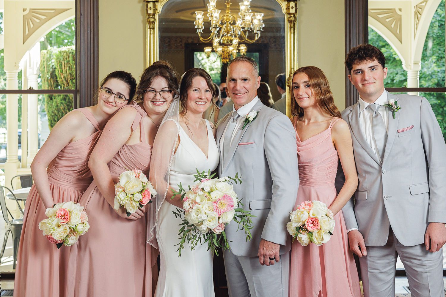 Bridal party stands in a line wearing blush pink dresses and light grey suits under an ornate chandelier.