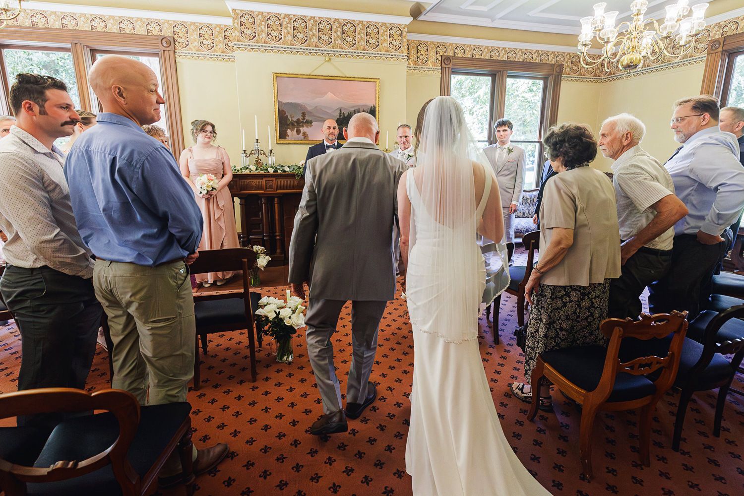 Guests gather during a wedding ceremony in an elegant room with ornate trim and patterned carpet.
