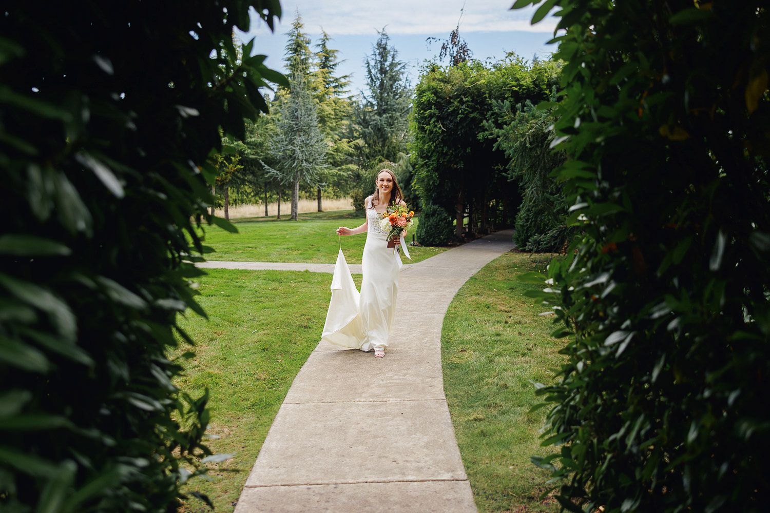 A person in a flowing white dress walks along a winding garden path surrounded by lush greenery on a sunny day.