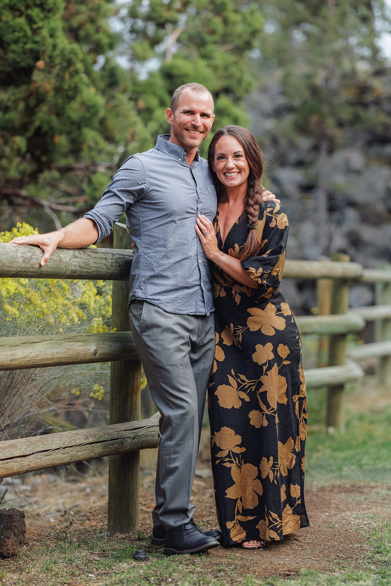 A couple in casual attire poses together against a wooden fence in a natural outdoor setting with greenery.
