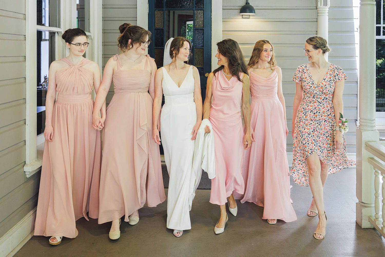 A group of bridesmaids in pink dresses pose together in a hallway with white walls and wooden floors.