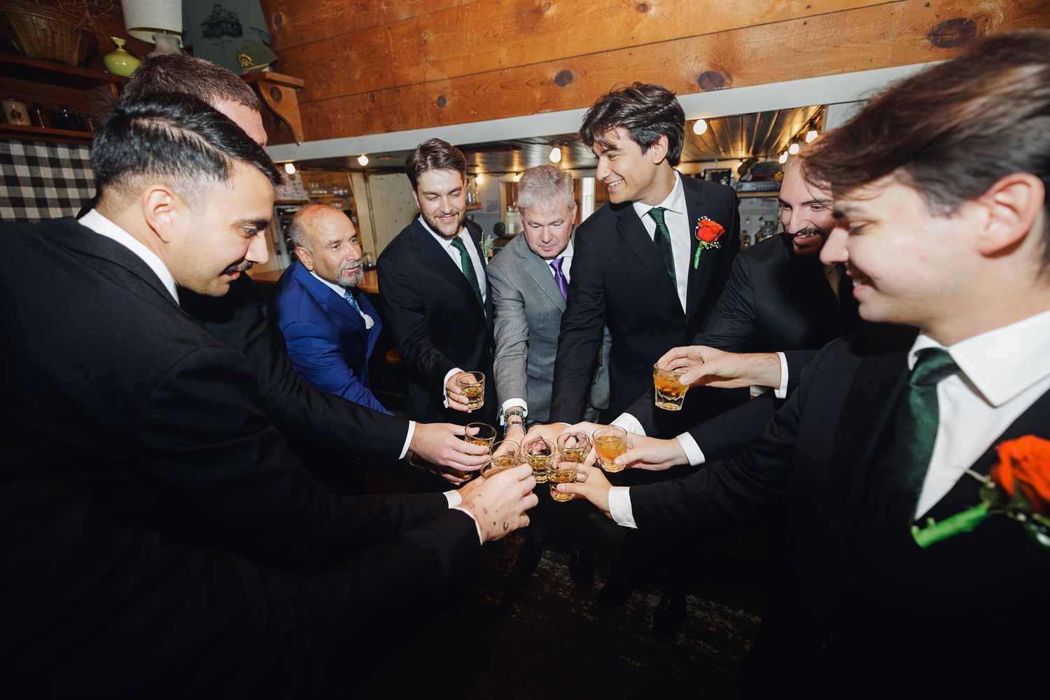 Group of groomsmen in suits toasting with drinks at a wedding reception in a rustic venue.