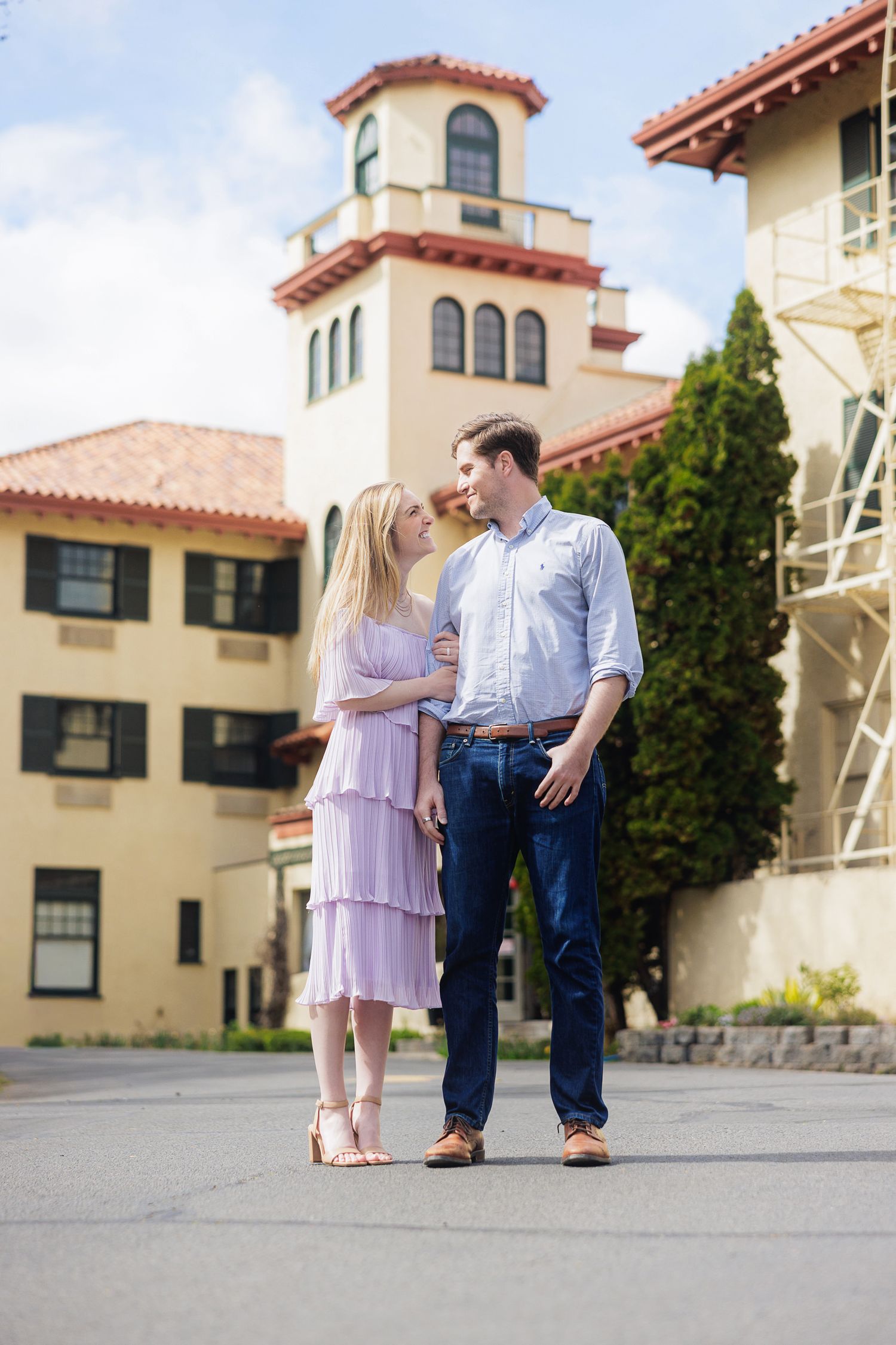 A couple in elegant attire stands together outside a Spanish-style building, gazing at each other romantically.