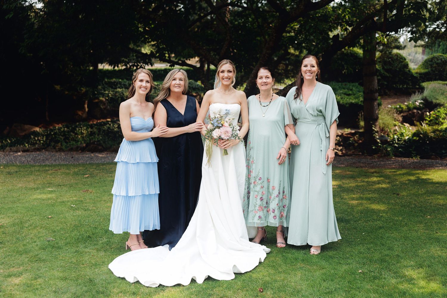 Bride in white gown poses with four women in blue and green dresses on a lush garden lawn.
