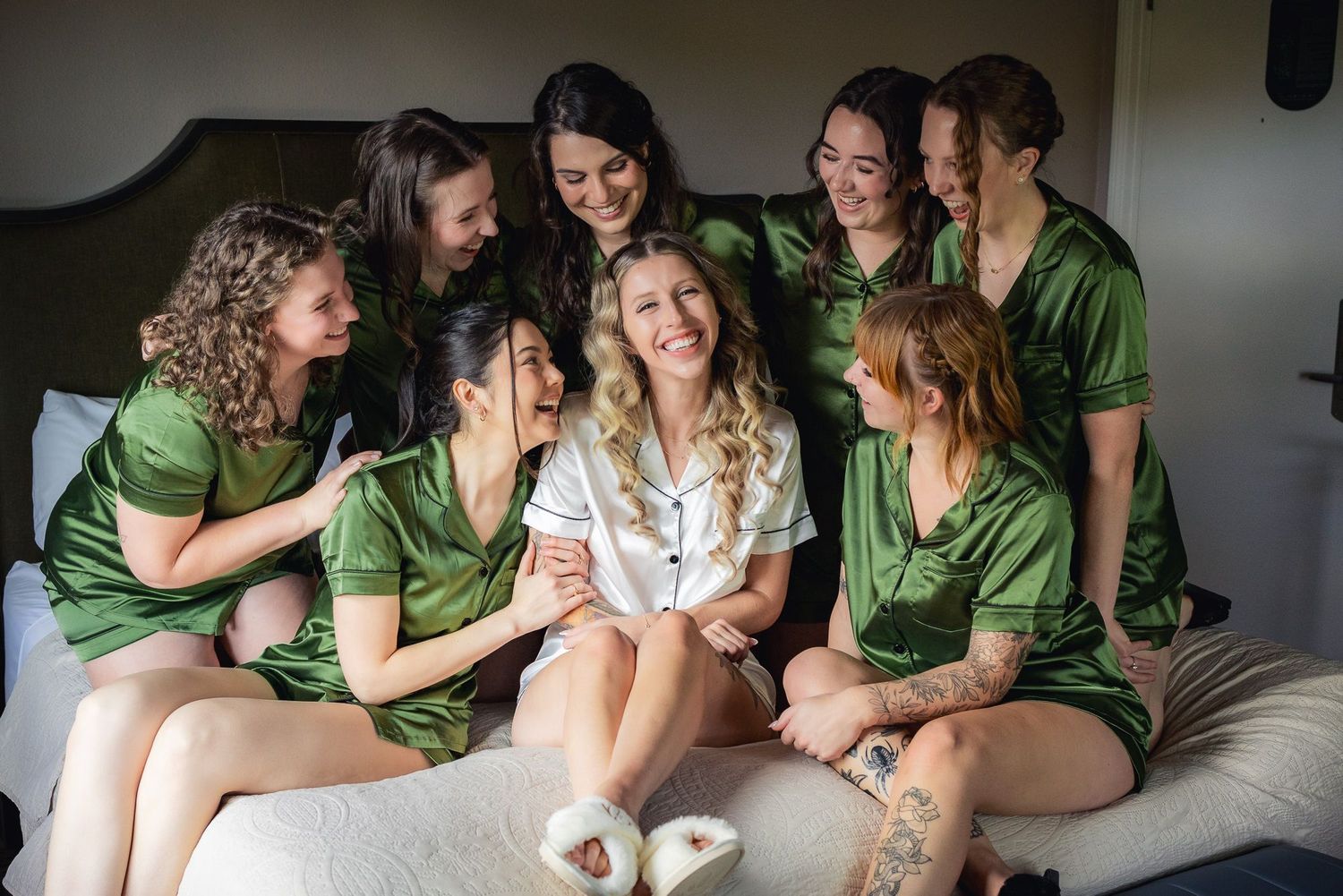 A group of bridesmaids wearing matching green robes gather around a bride in white on a couch sharing a joyful moment.