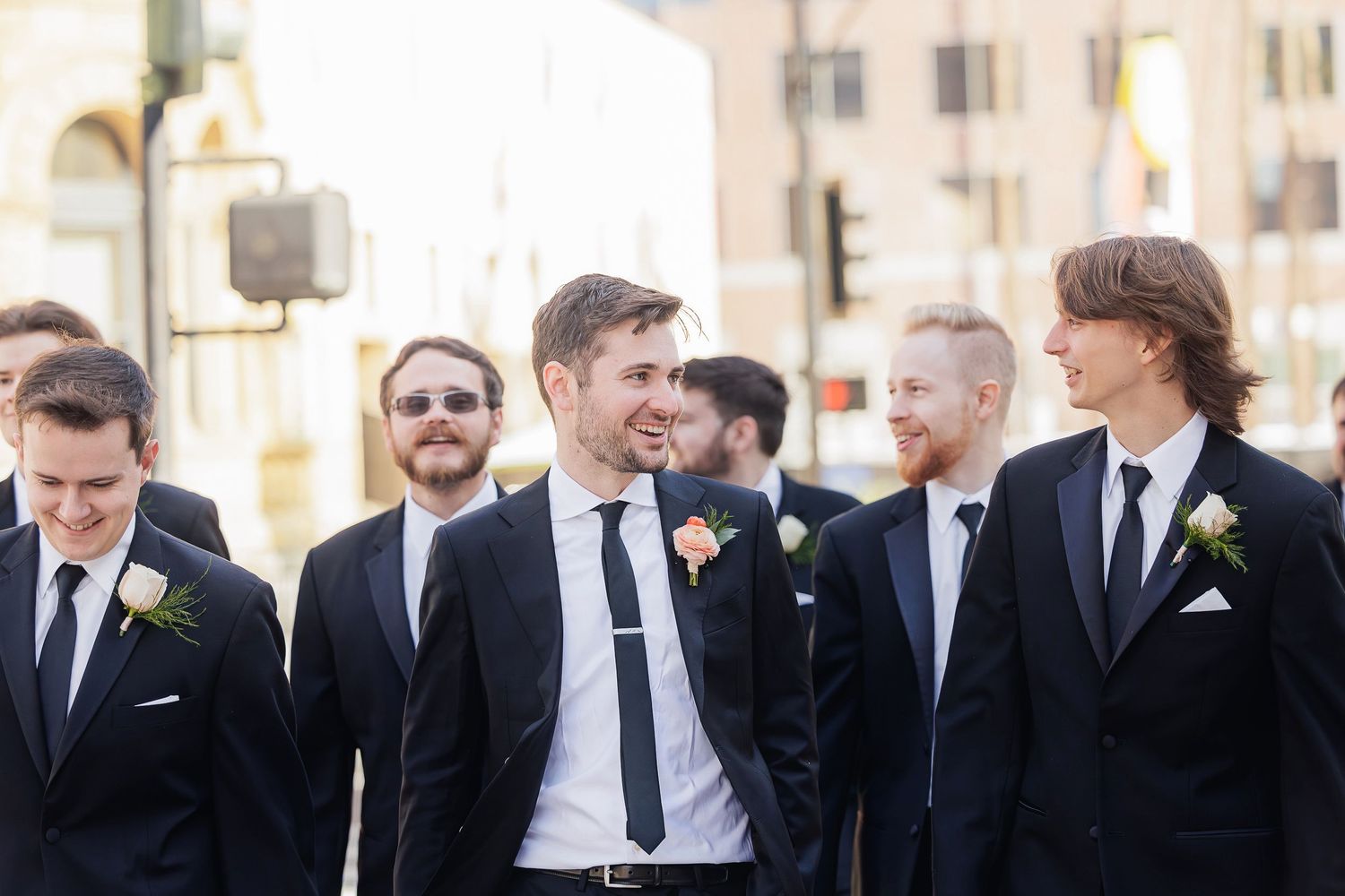Group of groomsmen in dark suits with boutonnieres smiling and laughing together outdoors.
