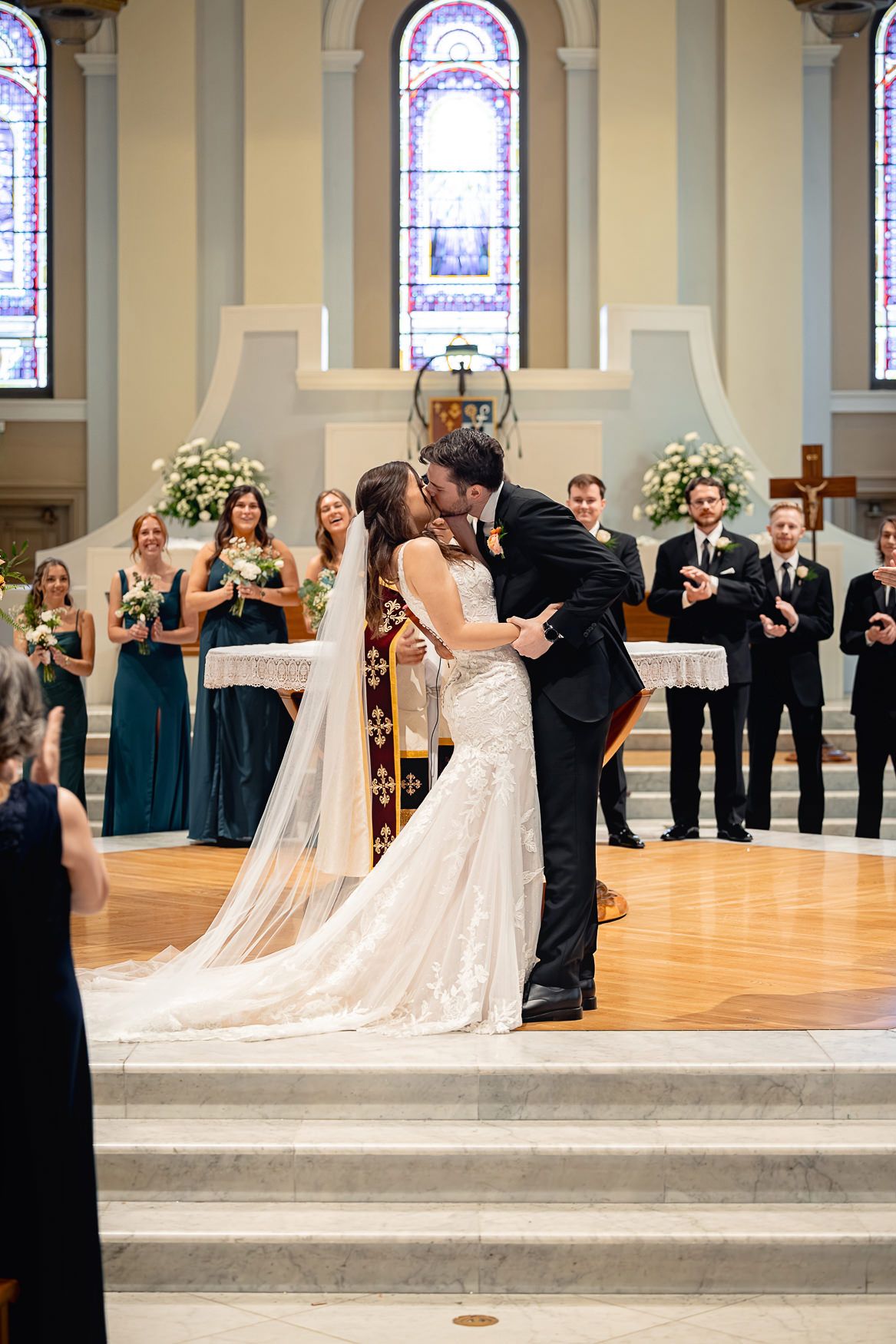 Wedding ceremony in a church with stained glass windows, bridesmaids in teal dresses, and floral arrangements at the altar.