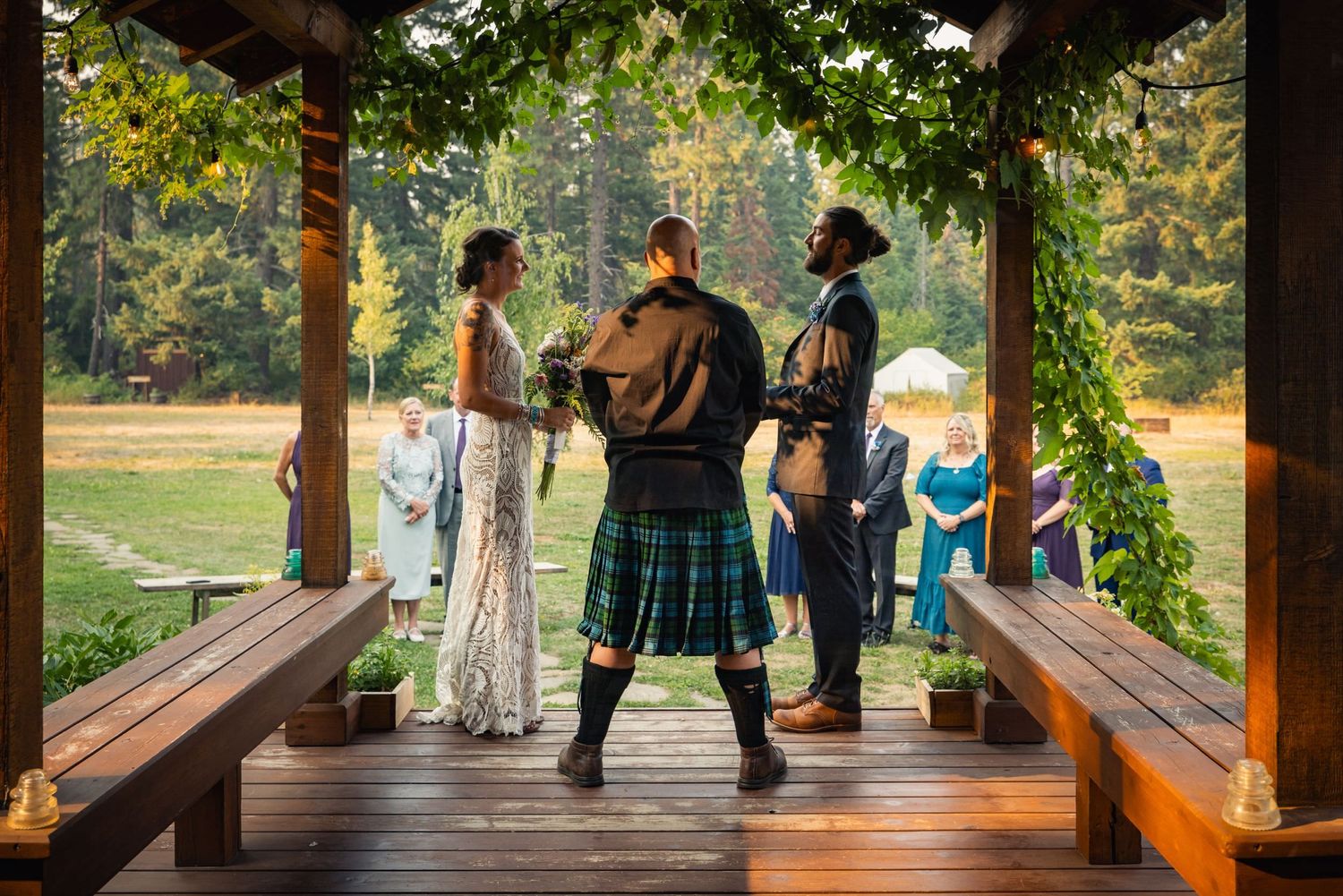 People gather on a rustic wooden porch surrounded by greenery while a bagpiper in traditional kilt performs.