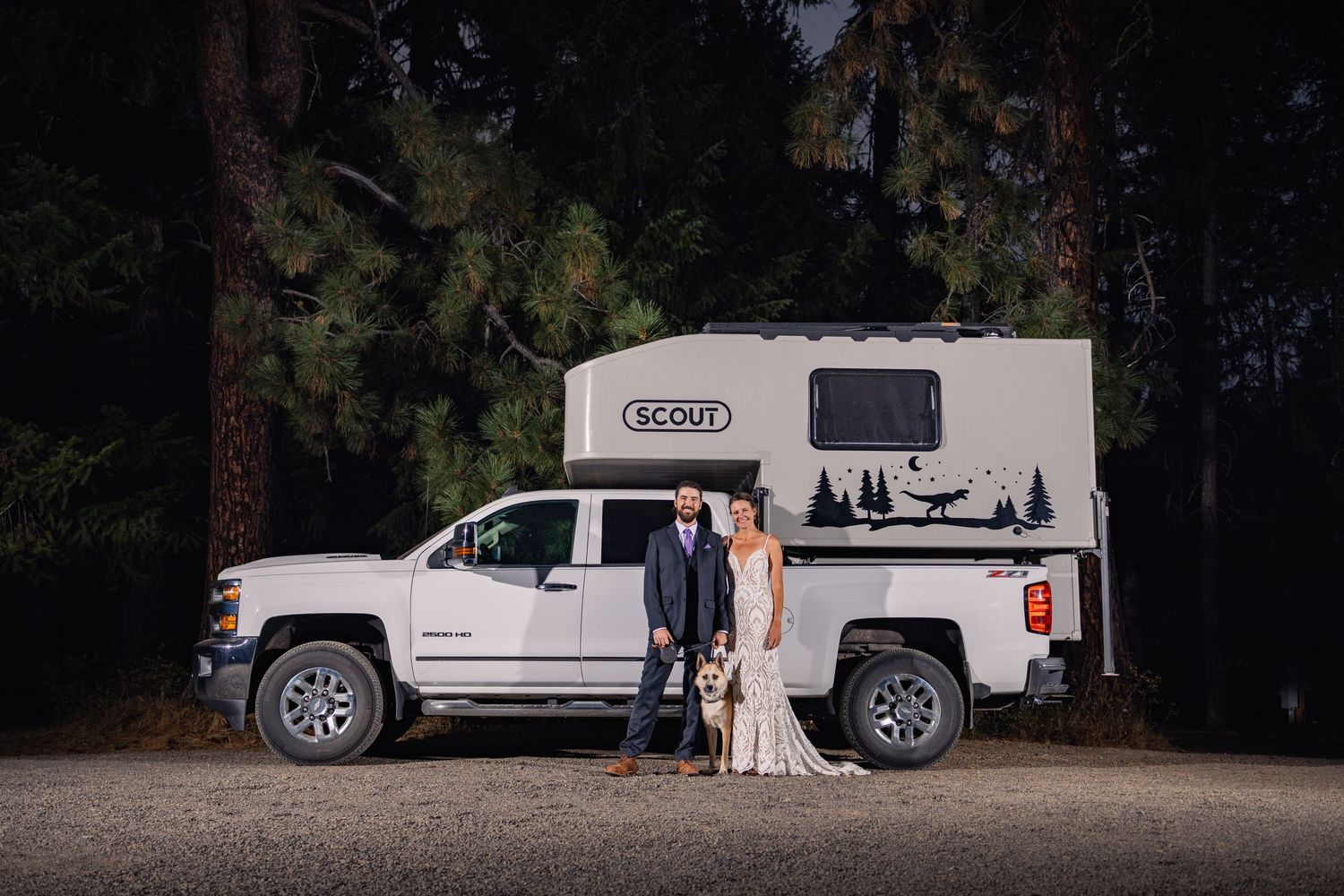 A couple in formal attire poses with their white pickup truck and camper in a forest setting at dusk.