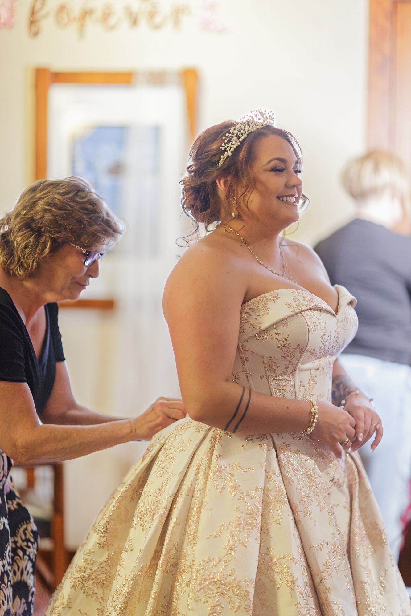 A bride wearing a champagne-colored ball gown with delicate embroidery and a crystal tiara smiles during wedding preparations.