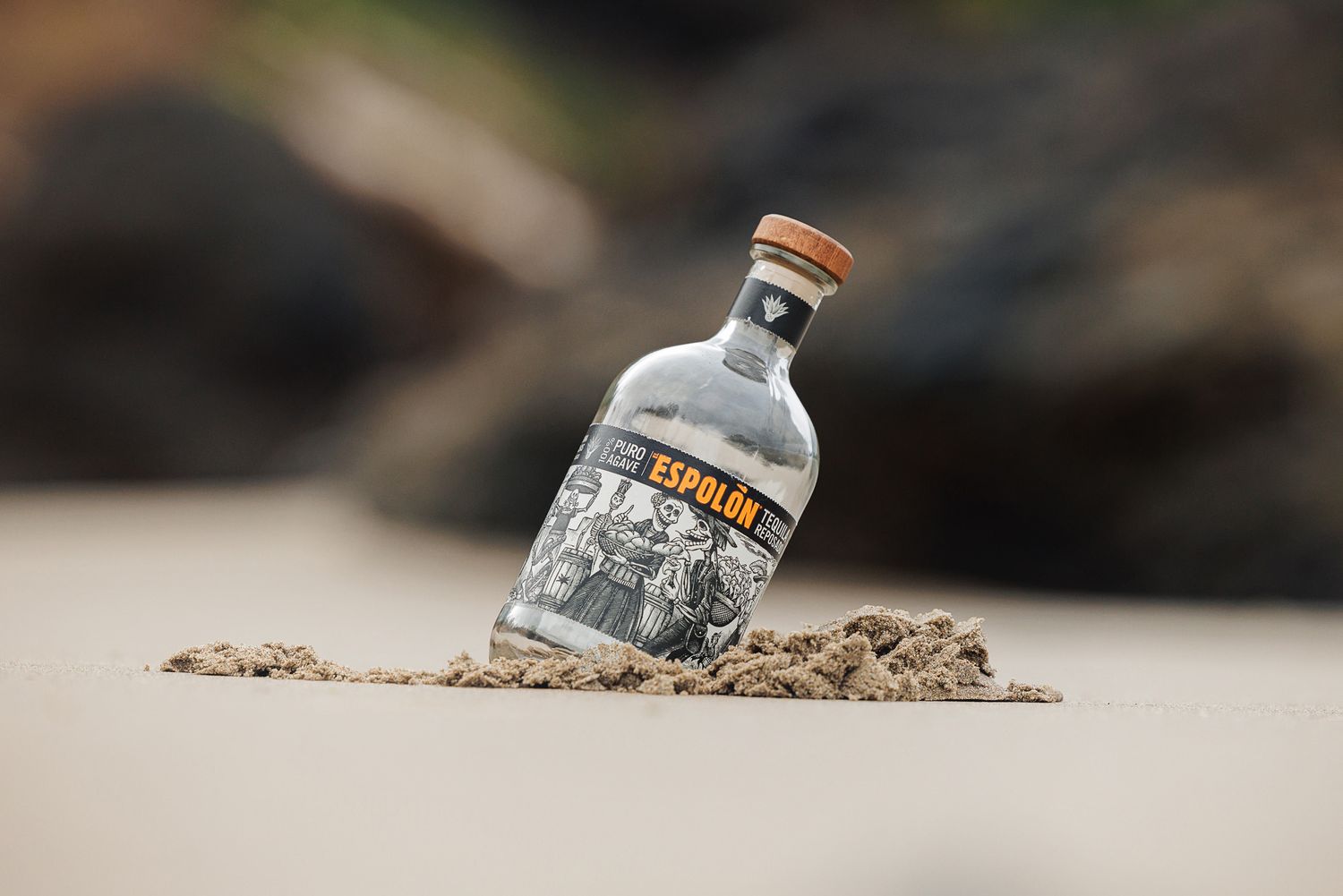 Espolon tequila bottle standing upright in sandy beach with blurred rocky background.