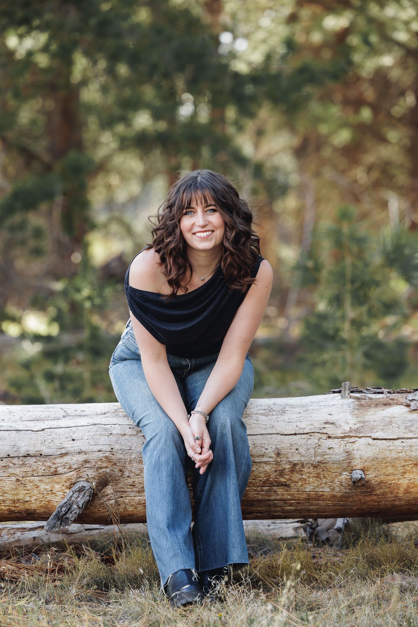 A smiling brunette woman sits on a log in a forest, wearing a black top and jeans, hands clasped.