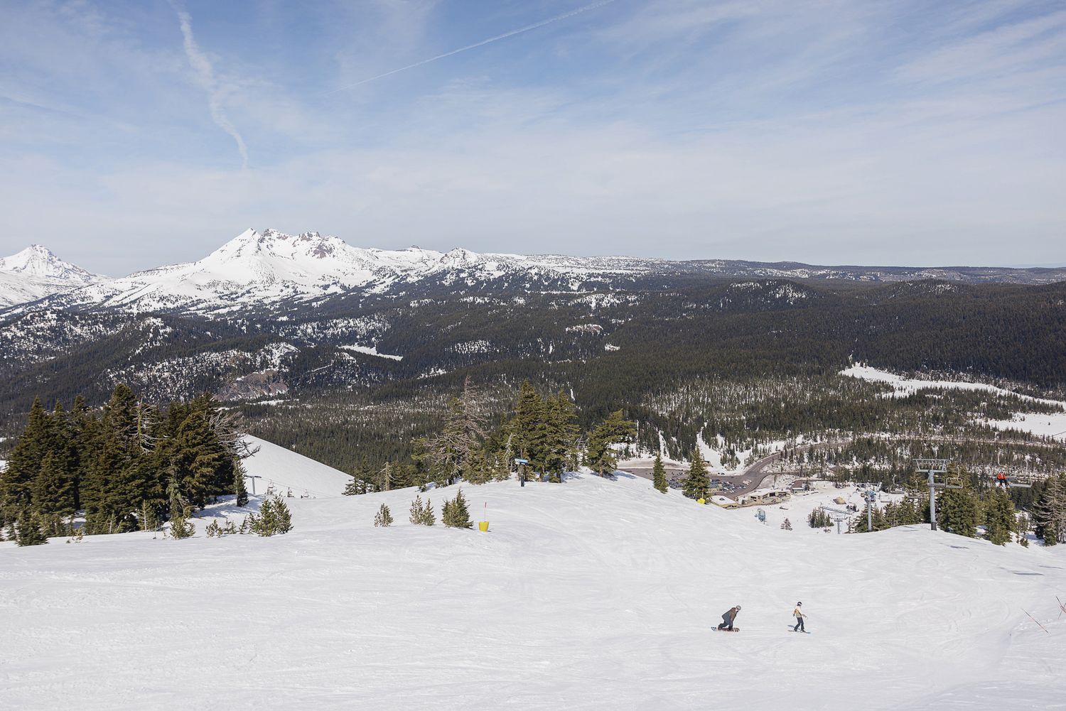 A snowy mountain landscape with skiers on the slopes, surrounded by pine trees and a cloudy sky.