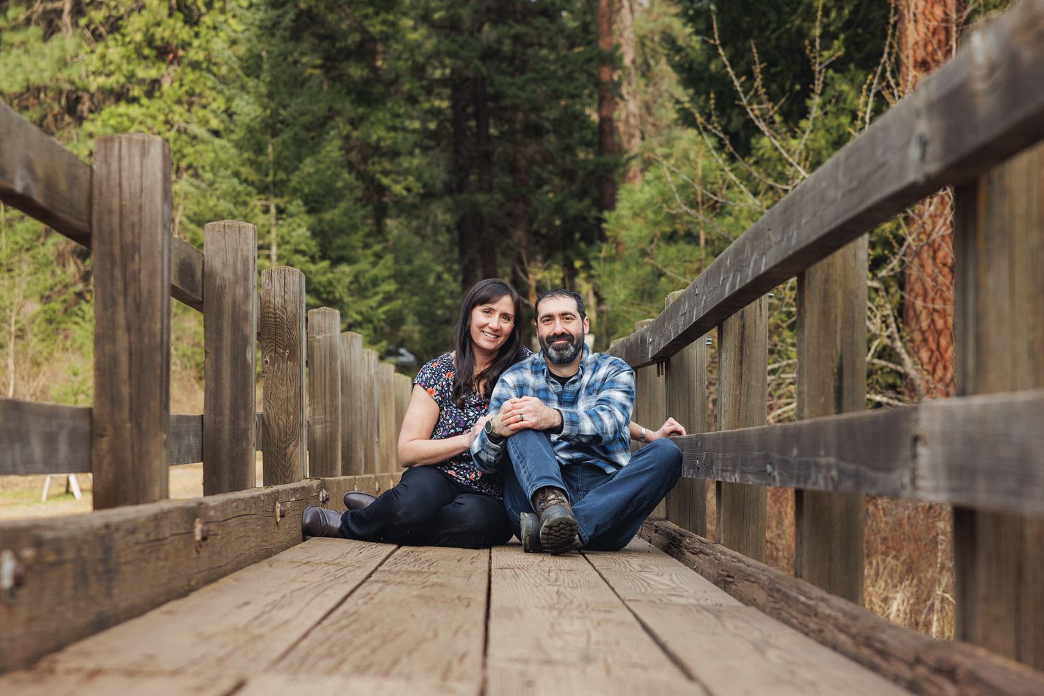 Couple sitting together on a wooden bridge surrounded by tall pine trees in a forest setting.