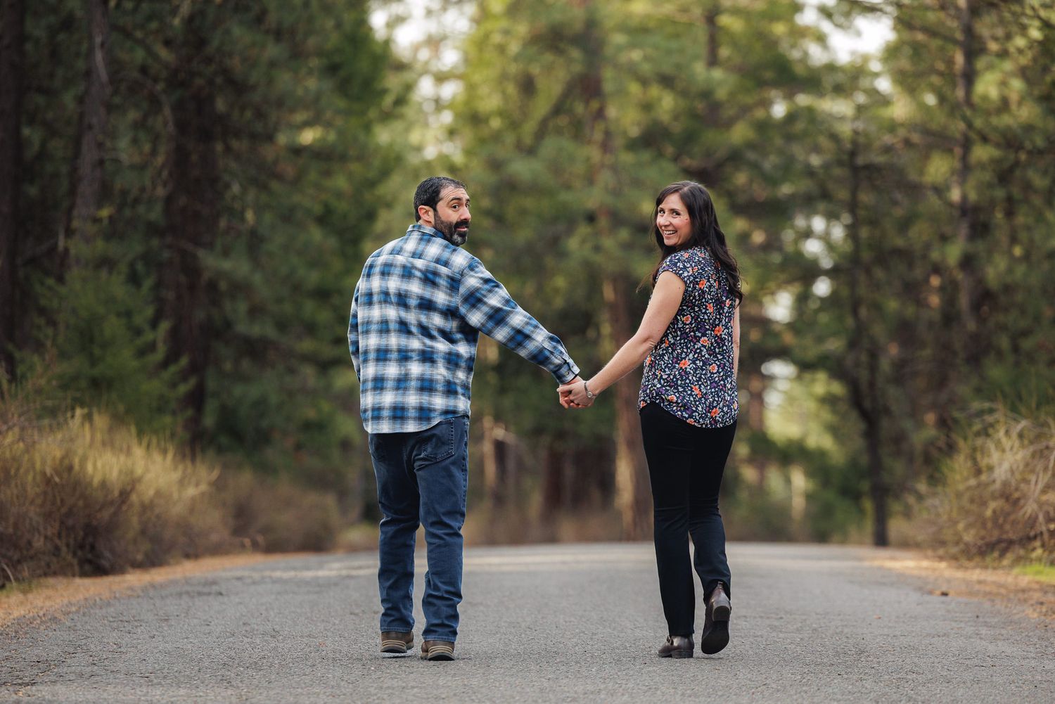 Couple holding hands on a tree-lined road, man in plaid shirt and woman in floral top, looking back smiling.