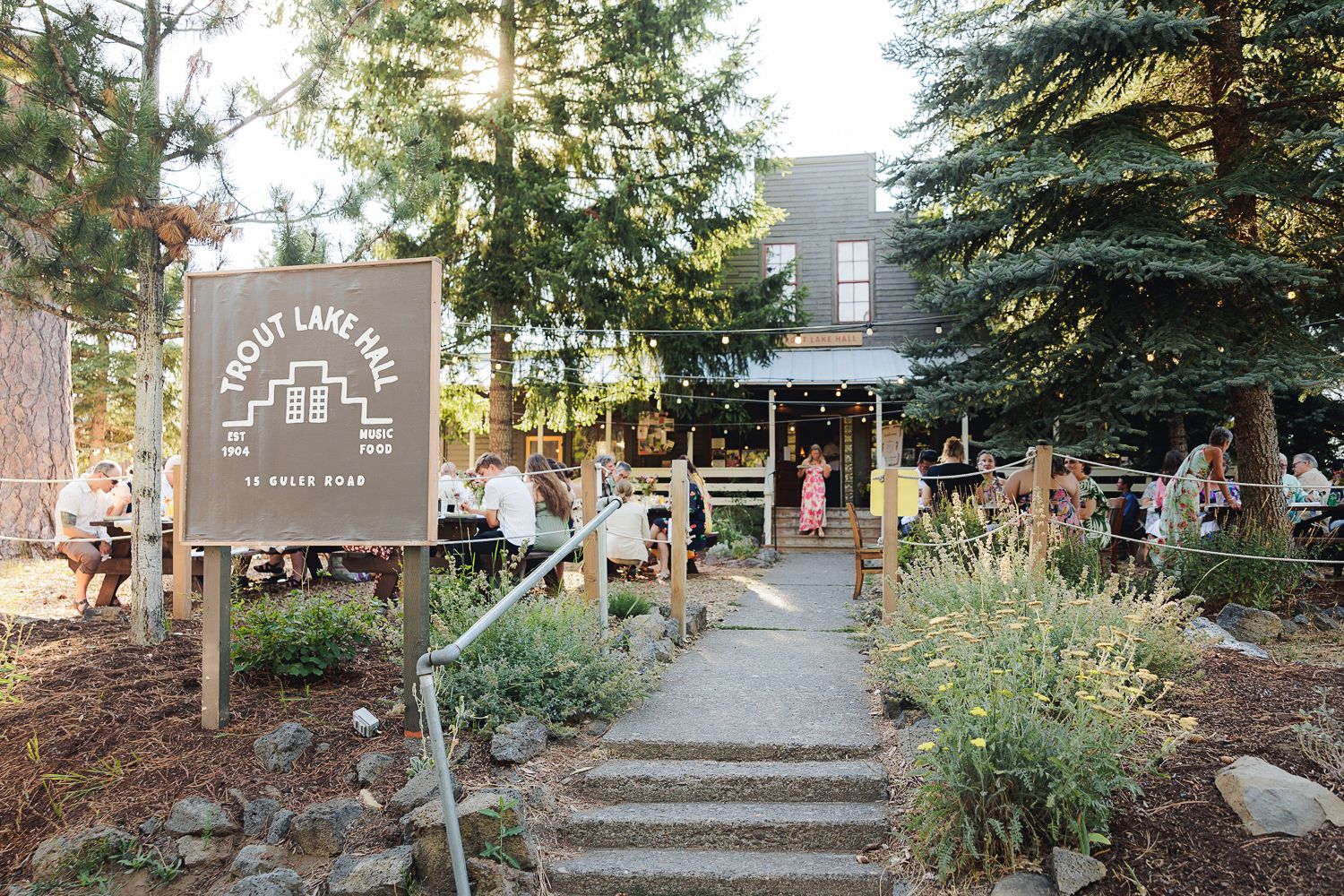 Garden pathway surrounded by evergreen trees and native plants leading to a rustic building entrance at dusk.