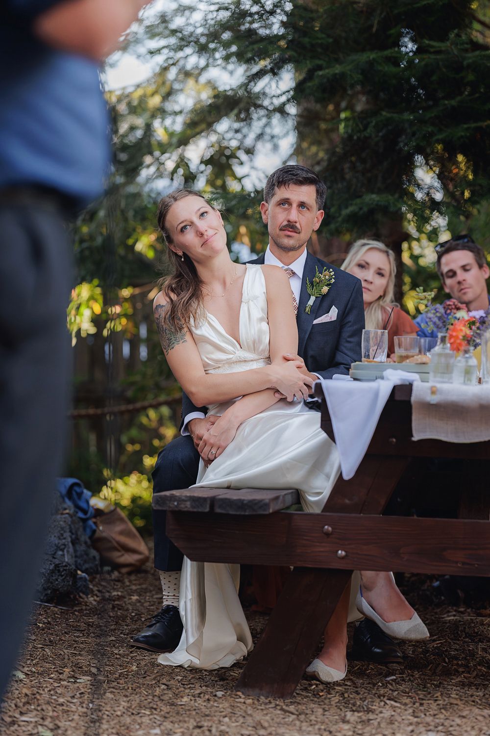 Newlywed couple sharing intimate moments while sitting on wooden benches during an outdoor reception at sunset.