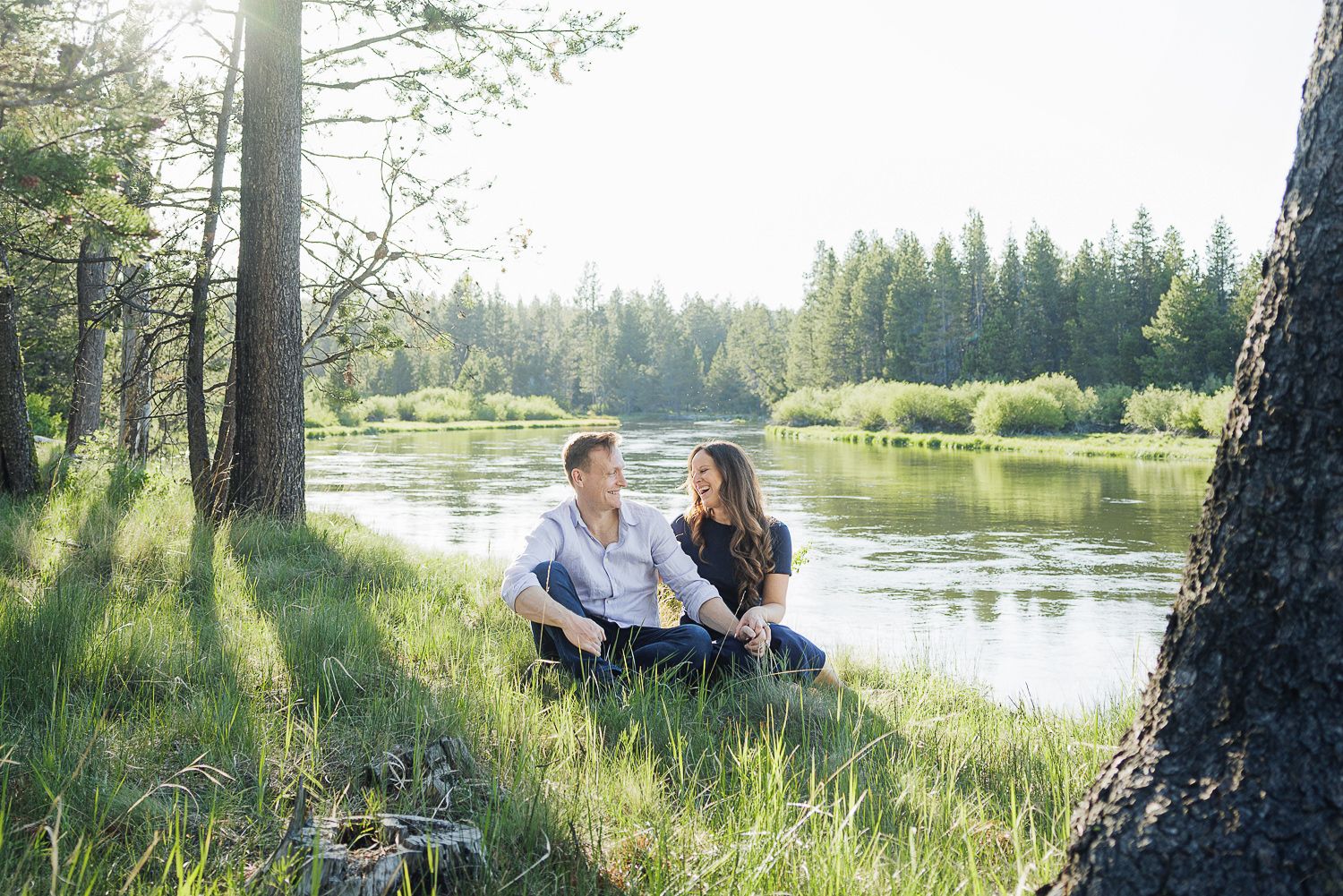 A couple sits together on grassy riverbank in sunlit forest setting beside tranquil water.