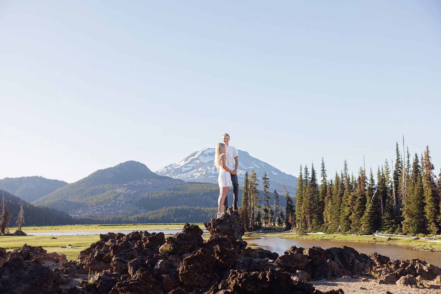 Couple standing on volcanic rocks with a snow-capped mountain and pine trees in the background on a clear day.