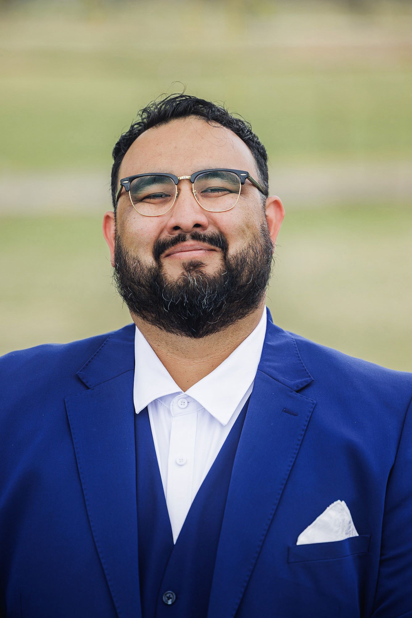 A bearded man wearing glasses and a blue suit with white pocket square smiles outdoors.