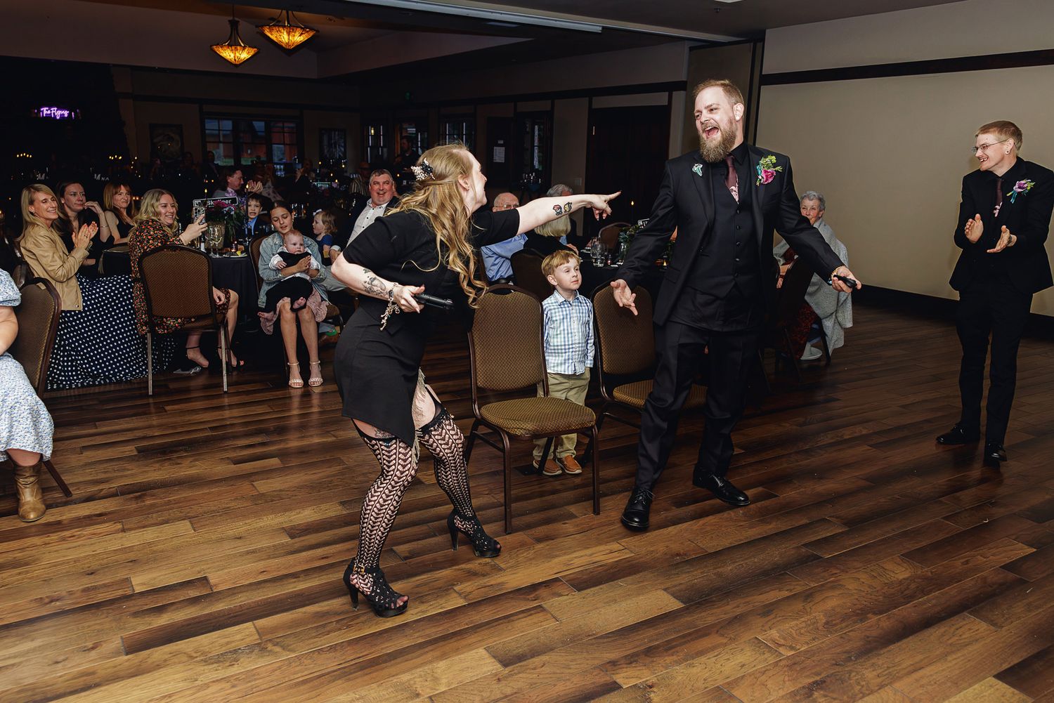 Couple dancing energetically on wooden dance floor at formal event while guests watch and applaud in dimly lit venue.