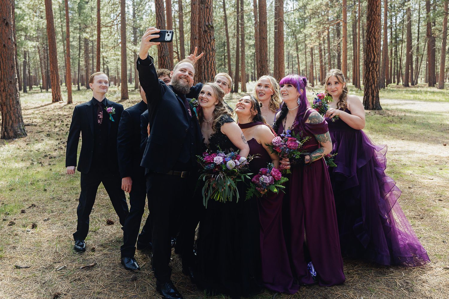Wedding party in purple gowns and black suits taking a selfie among tall pine trees in a forest setting.