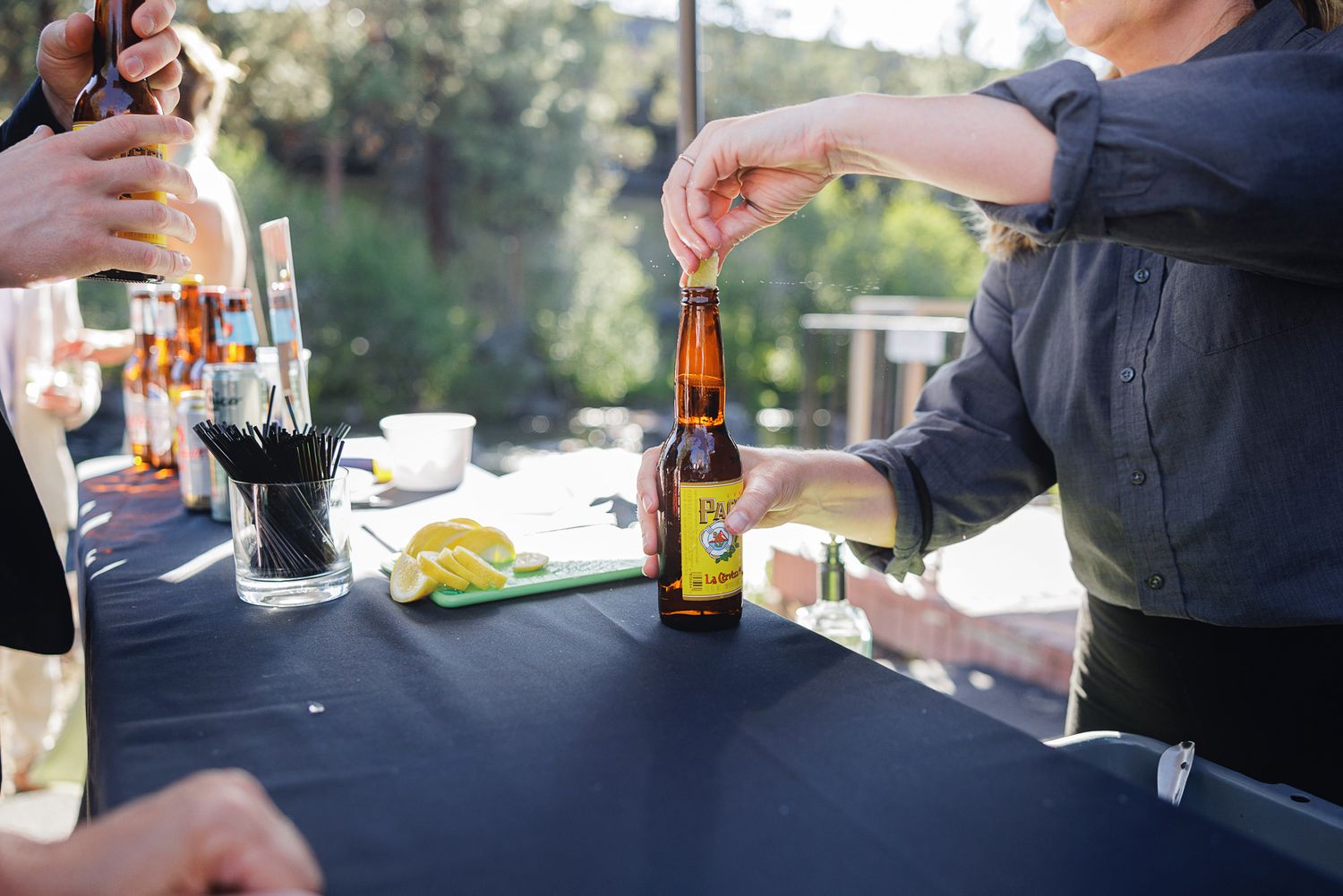 Person opening a craft beer bottle at an outdoor bar table with limes and drinks on a black tablecloth.