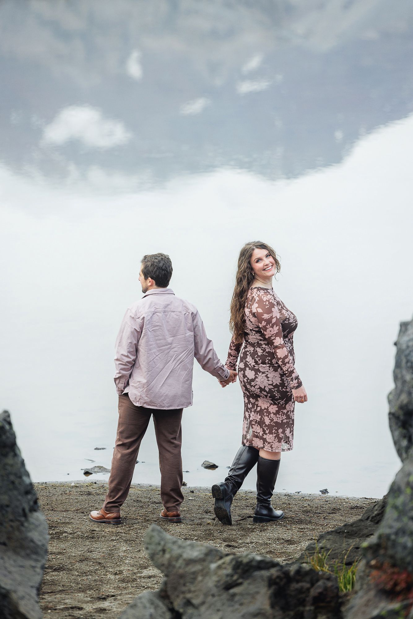 A couple in coordinating outfits stands together on a rocky cliff overlooking a cloudy landscape.