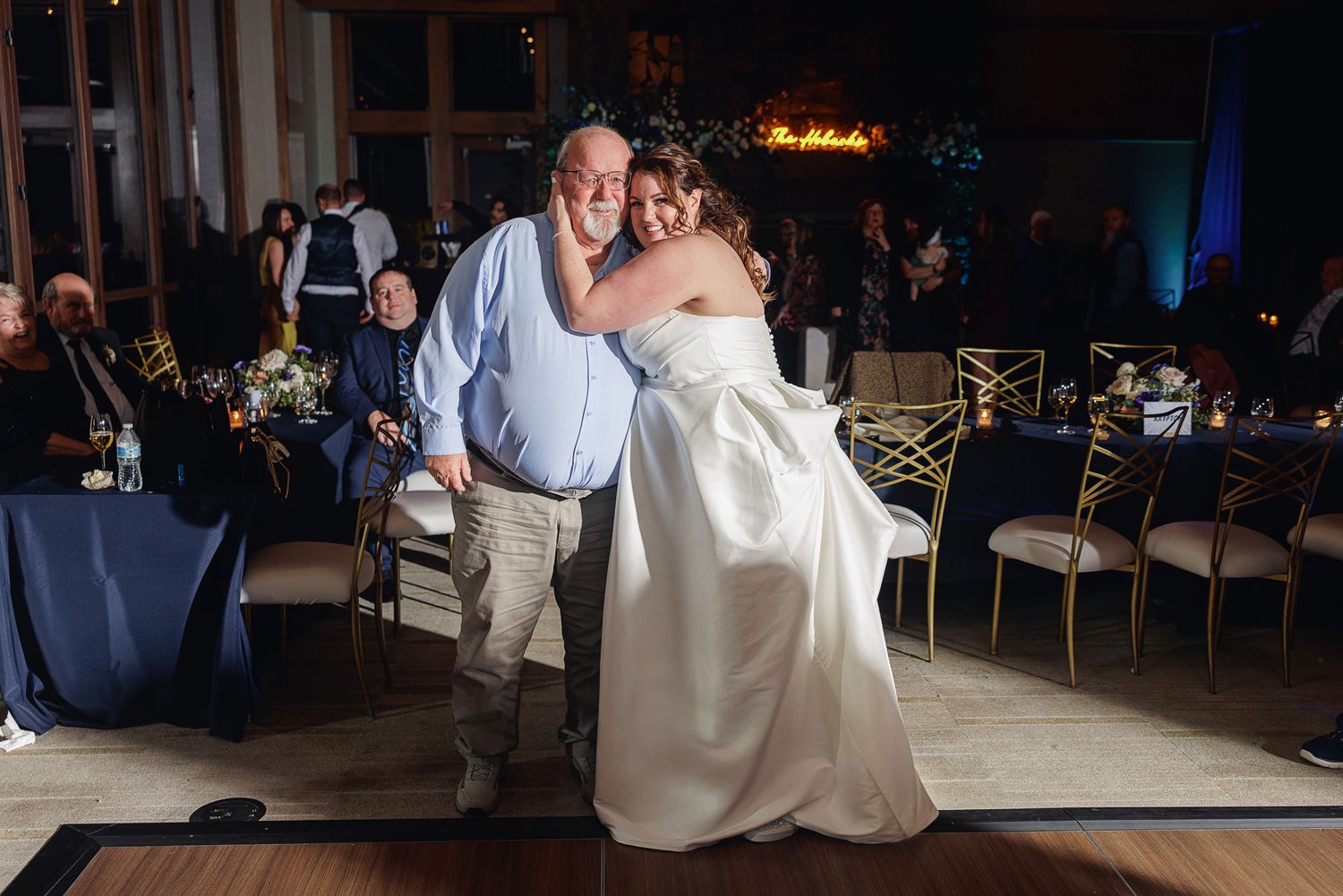 Wedding guests dance together during an evening reception on a dark dance floor with blue lighting.