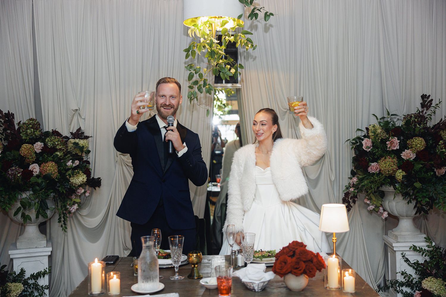 Bride and groom raising glasses in a toast at elegant wedding reception with floral arrangements and candles.
