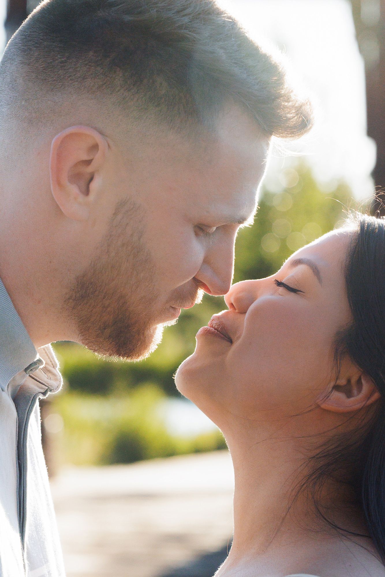 A romantic couple sharing an intimate moment, their faces close together about to kiss outdoors in soft sunlight.