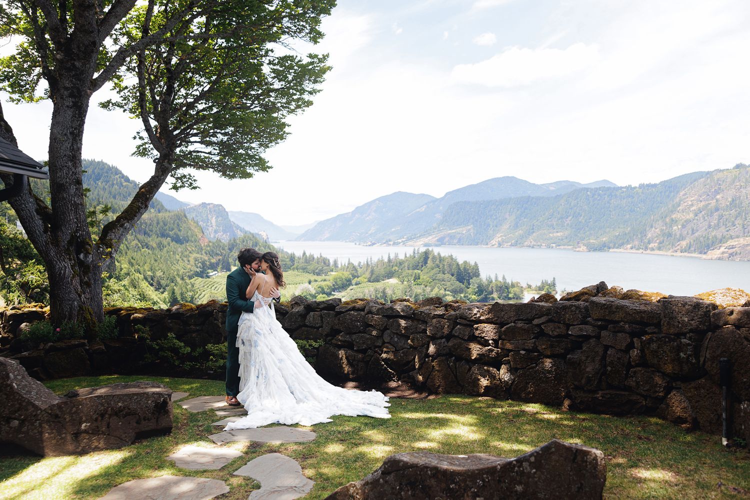 Bride and groom embrace at stone wall overlook with Columbia River Gorge mountains and water views on their wedding day.