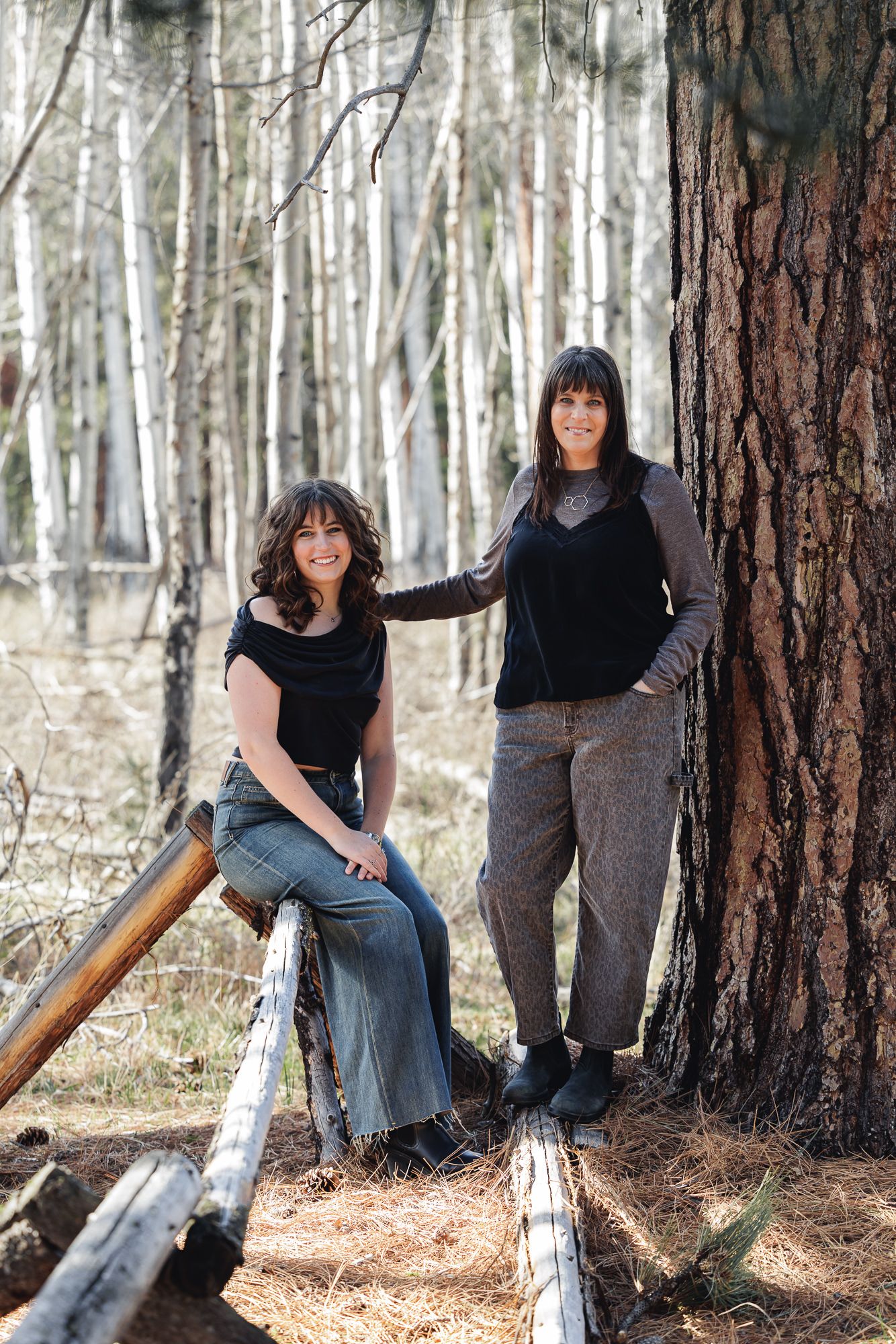 Two brunette women posing in a forest, one sitting on a log and one leaning against a pine tree.