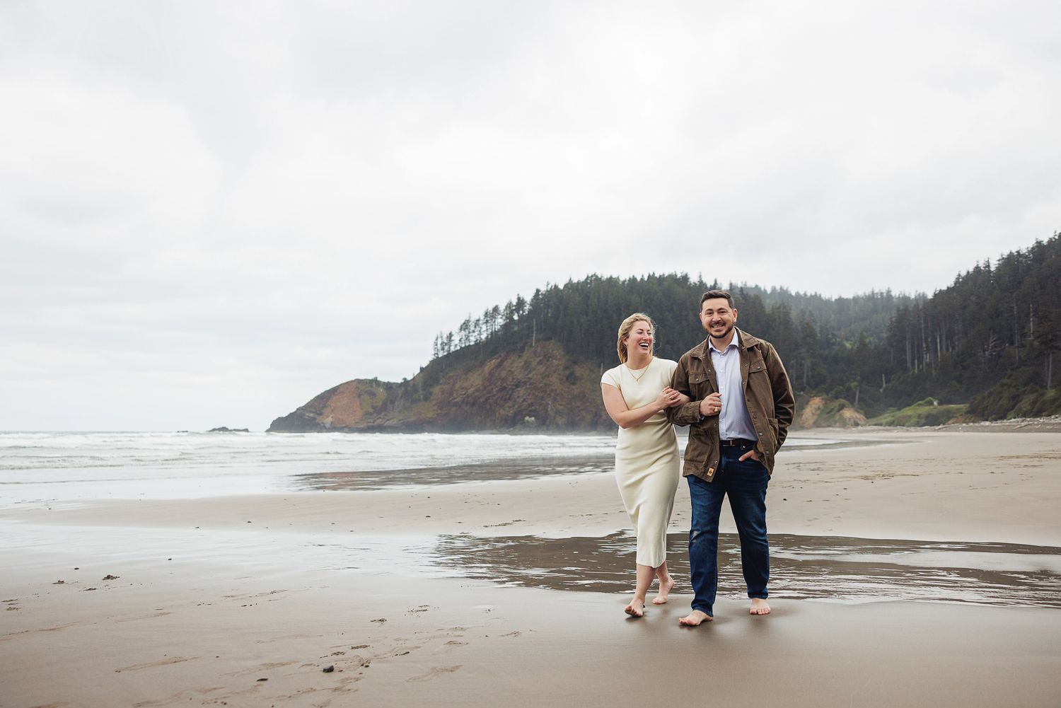 Couple smiling and walking hand in hand on a sandy Oregon beach with misty cliffs and evergreen trees in background.