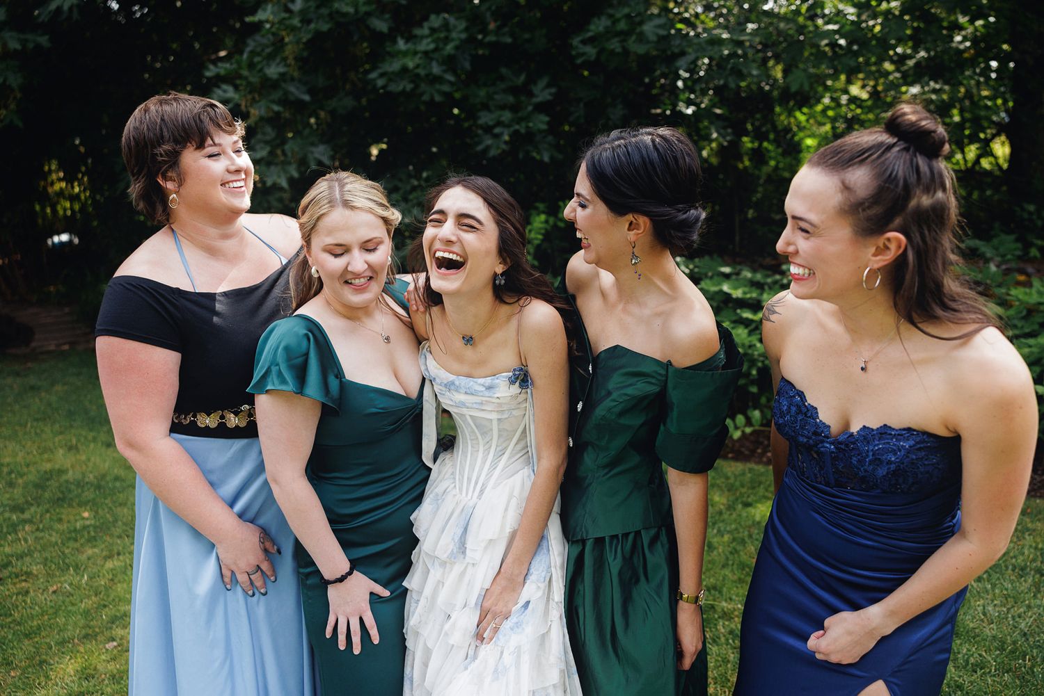 Five women in formal gowns laughing together outdoors at a wedding celebration.