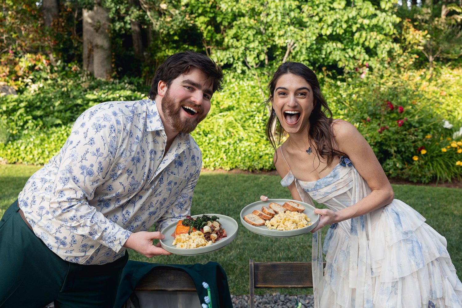 Couple smiling and holding plates of food outdoors in a lush garden setting at a wedding reception.