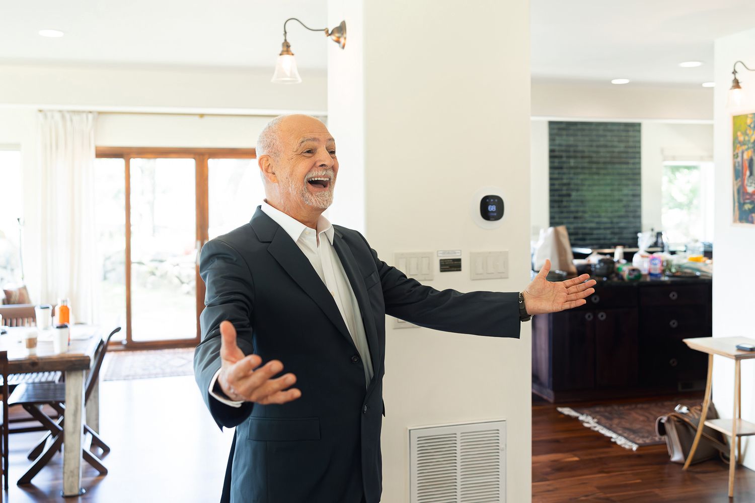 Bald man in suit gesturing welcomingly inside a modern bright home with smart thermostat on white wall.
